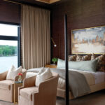 Dark wood wall paneling, beige upholstered chairs, and a four-poster bed in a bedroom.