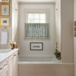 White tile bathroom with window, gingham blind, and framed artwork.