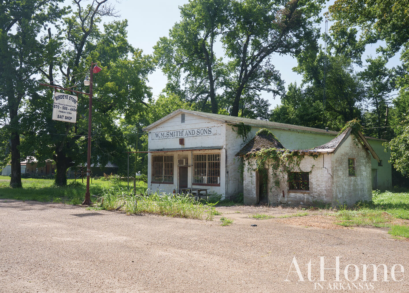 Small white building with a sign, surrounded by trees and a road.