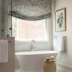 Gray patterned roman shade, marble bathtub, and brick wall in bathroom.