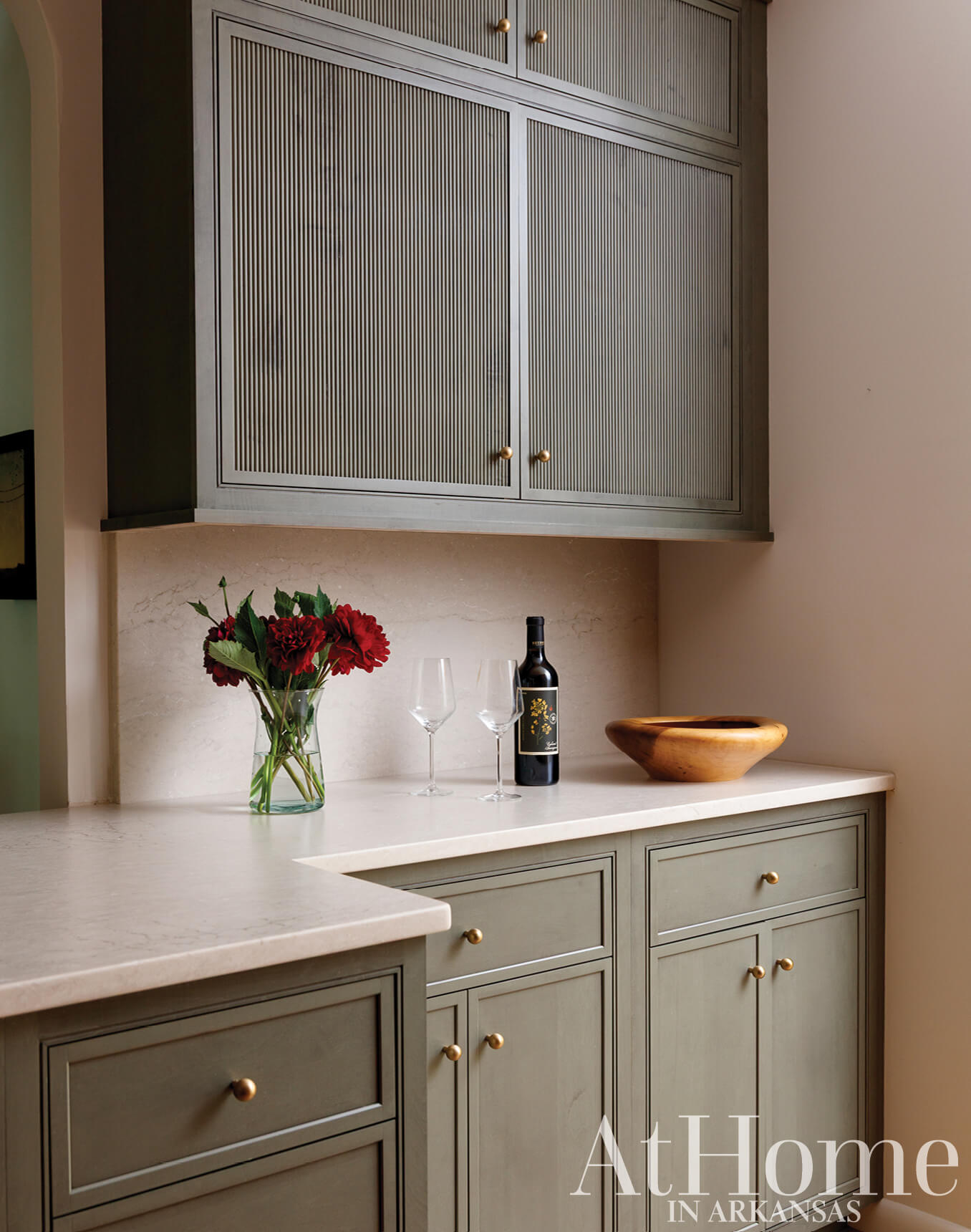 Gray and green cabinetry with brass hardware, wine, and flowers.