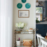 Neutral dining room with blue-glazed ceramic plates, framed artwork, and a bar cart.