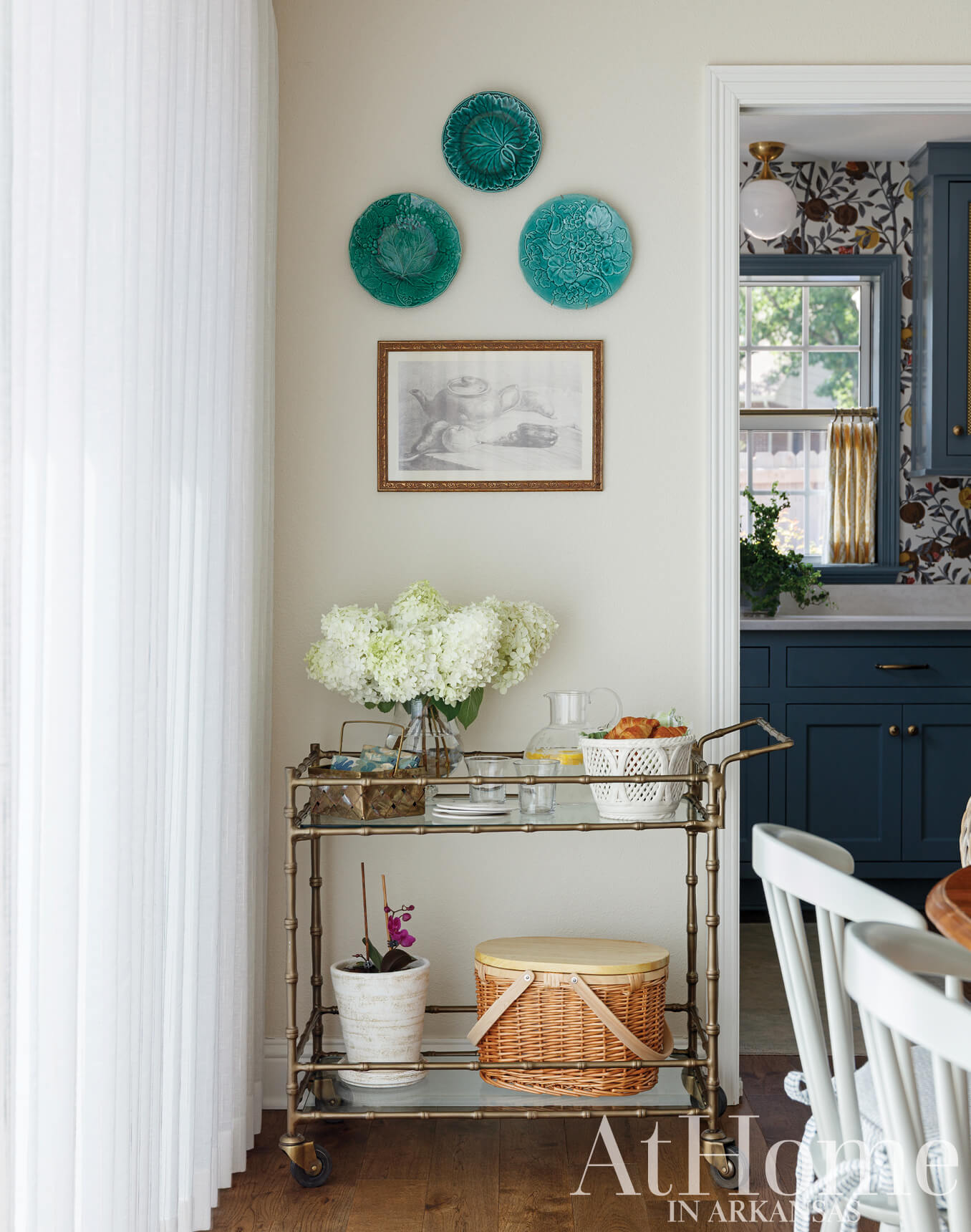 Neutral dining room with blue-glazed ceramic plates, framed artwork, and a bar cart.