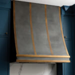 Dark metal range hood over teal cabinetry and marble countertop.